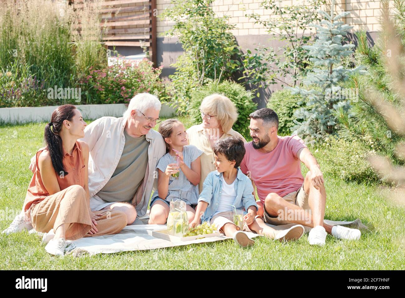 Ritratto di gioiosa famiglia multigenerativa seduta sul prato in cortile nella soleggiata giornata estiva con picnic Foto Stock