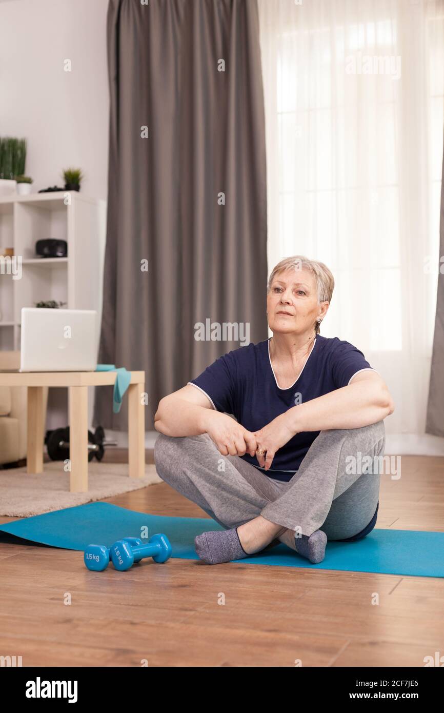 Donna seduta sul tappetino yoga in attesa dell'allenatore di benessere. Anziano pensionato online internet esercizio di formazione a casa attività sportiva con manubri, banda di resistenza, palla svizzera all'età di anziano pensionamento Foto Stock