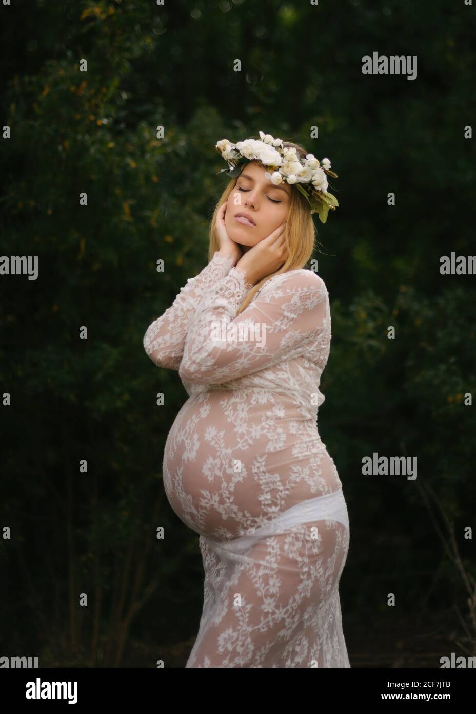 Calmo attraente donna incinta con occhi chiusi in pizzo bianco abito e bianco fiore corona sognare mentre in piedi e toccare testa tra le foglie di alberi verdi densi nella foresta in estate Foto Stock