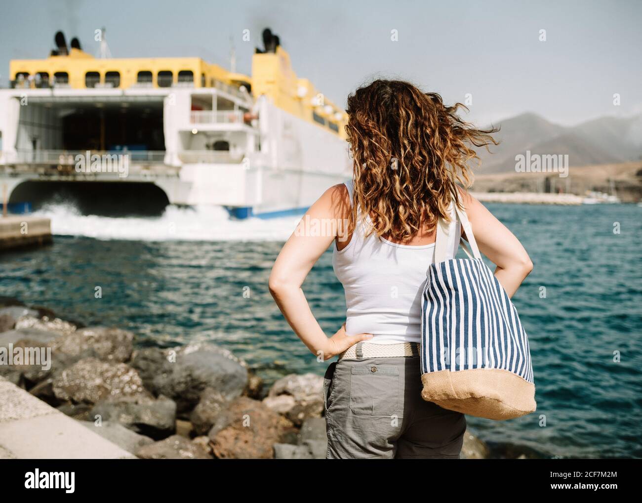 Vista posteriore della donna attiva con le mani a parte guardando la grande barca in onde turchesi sul molo a Fuerteventura, Las Palmas, Spagna Foto Stock