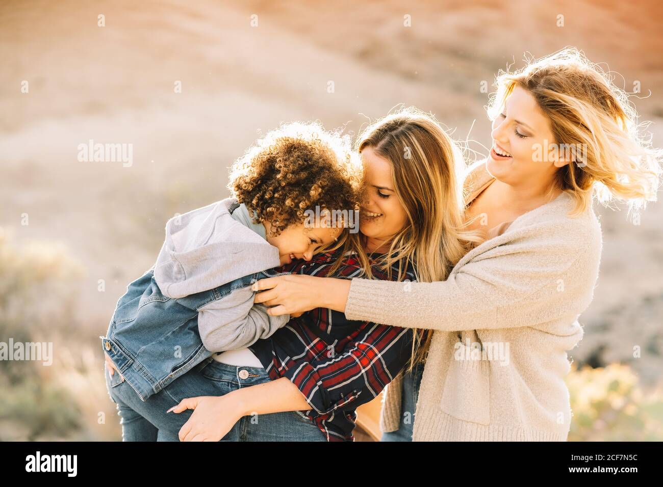 Donna allegra in camicia di controllo che tiene in braccio bambino casual con capelli ricci mentre amico femmina gioioso mettere cappello sul bambino sulla natura di giorno Foto Stock