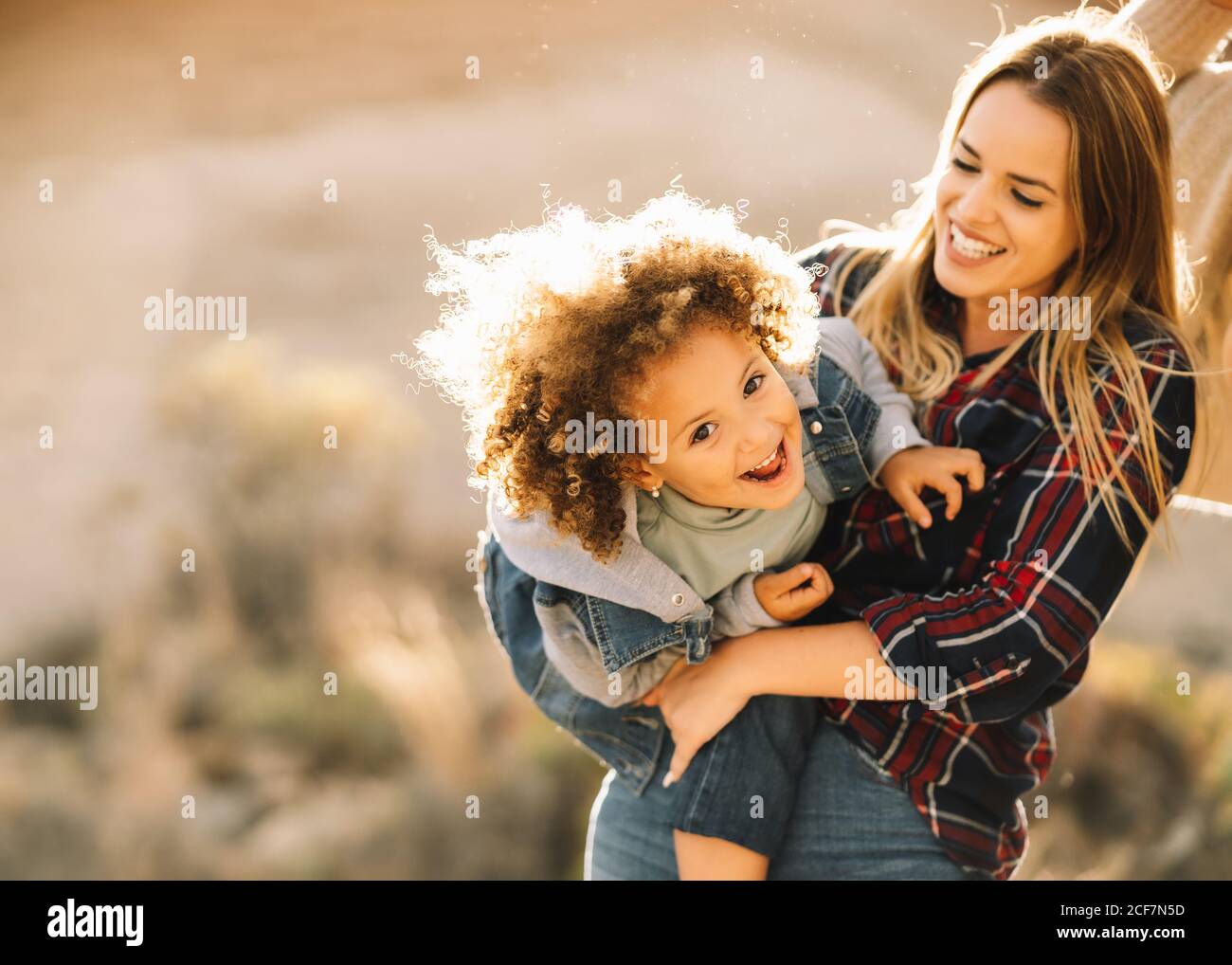 Gioiosa donna bionda in camicia di controllo che tiene le braccia coccolarsi bambino felice con i capelli ricci che guardano la macchina fotografica all'esterno a. giorno Foto Stock