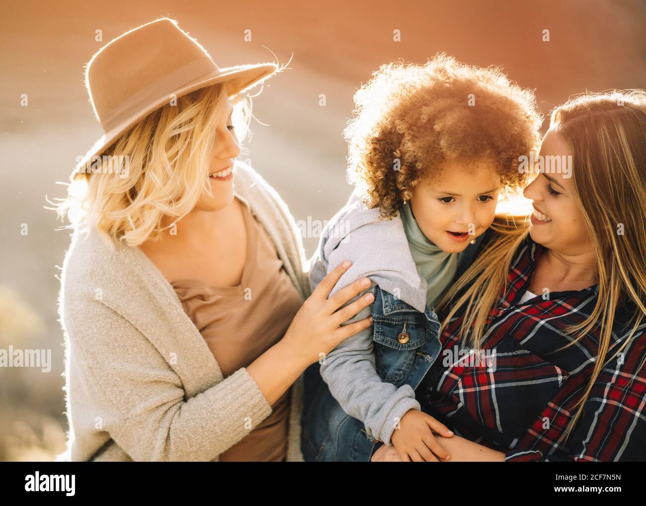Donna allegra in camicia di controllo che tiene in braccio bambino casual con capelli ricci mentre amico femmina gioioso mettere cappello sul bambino sulla natura di giorno Foto Stock