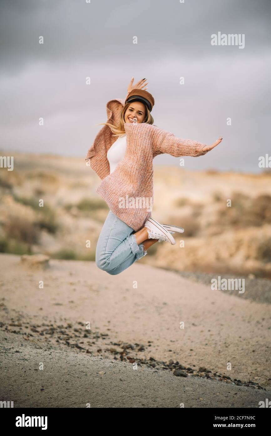 Femmina sorridente con capelli biondi vestiti eleganti e. cappello che salta con le braccia allungate e le gambe piegate terra sabbiosa e guardando la macchina fotografica Foto Stock