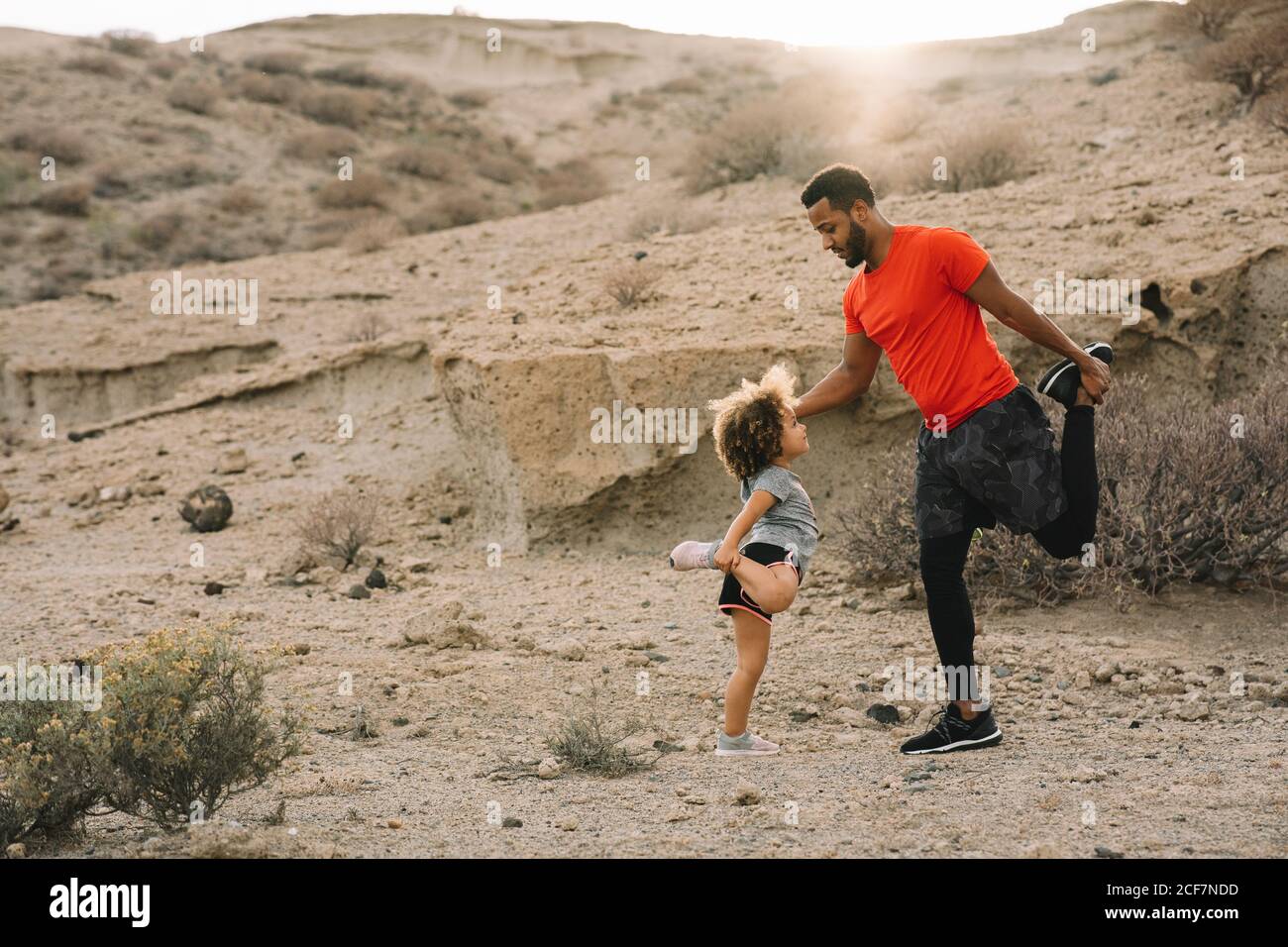 Vista laterale dell'uomo alato afroamericano in piedi activewear di fronte al bambino arricciato che insegna a stretching parte anteriore della coscia su sabbia orizzontale Foto Stock