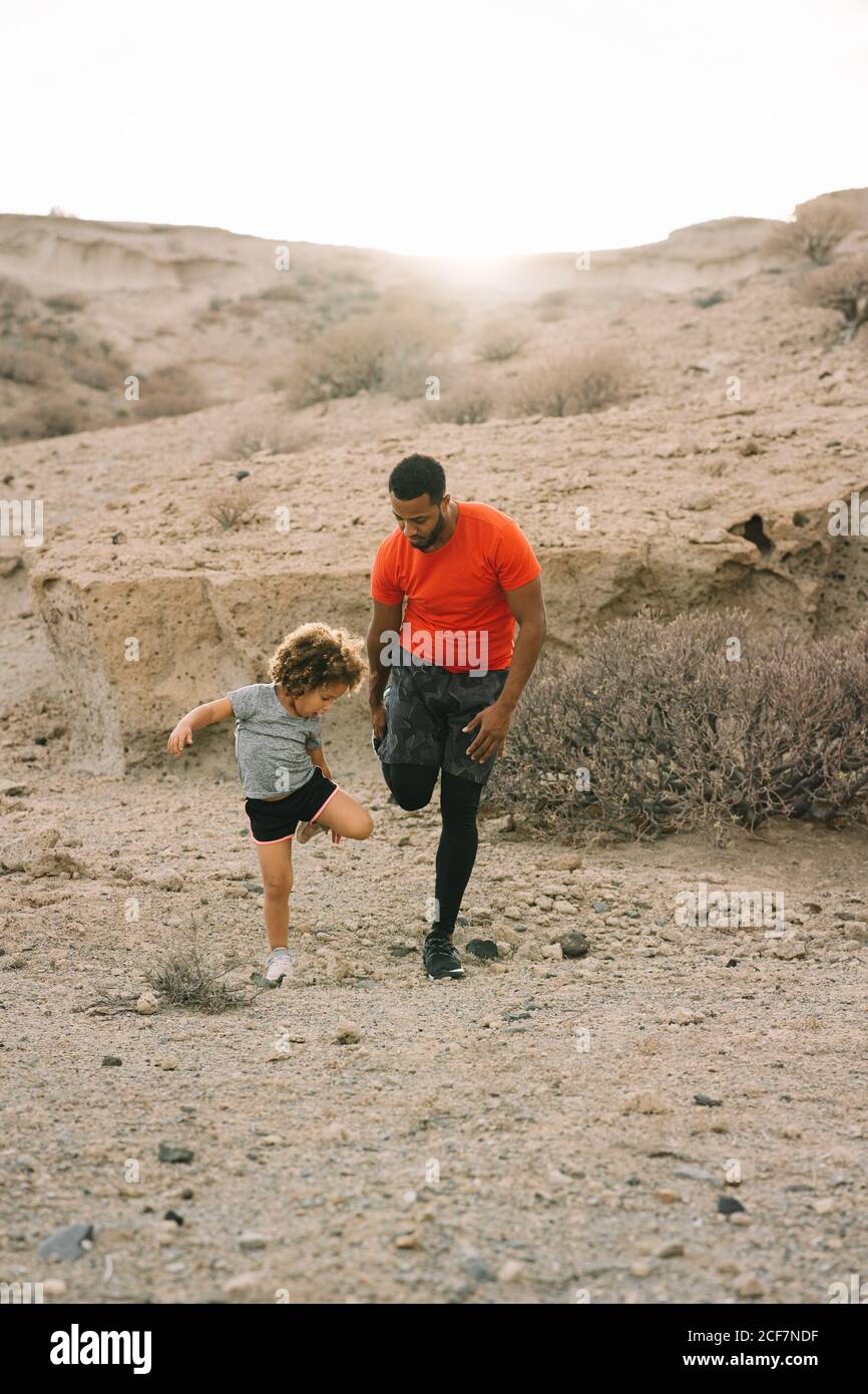 Vista laterale dell'uomo alato afroamericano in piedi activewear di fronte al bambino arricciato che insegna a stretching parte anteriore della coscia su sabbia orizzontale Foto Stock