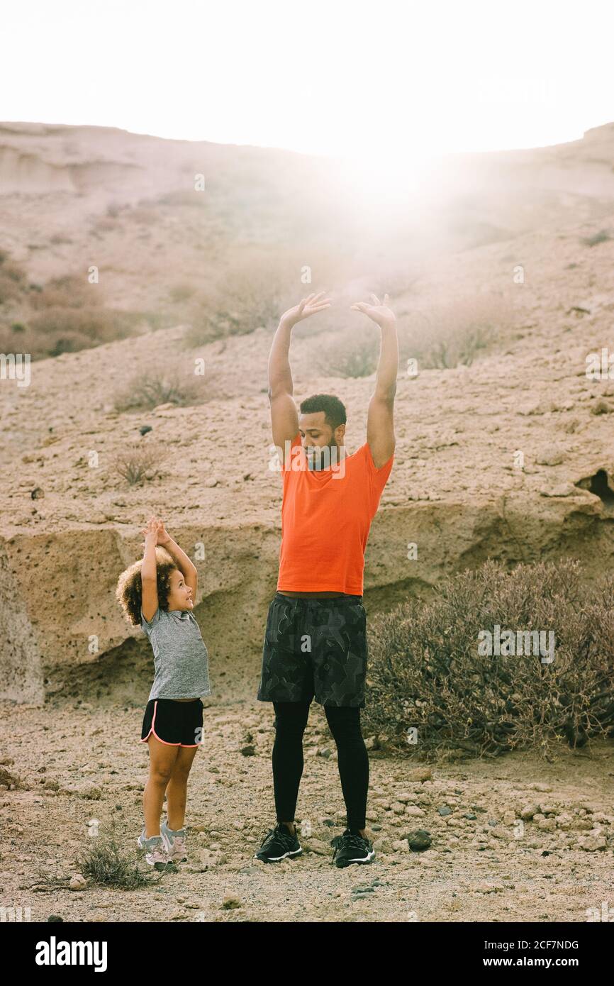 Vista laterale dell'uomo alato afroamericano in piedi activewear di fronte al bambino arricciato che insegna a stretching parte anteriore della coscia su sabbia orizzontale Foto Stock