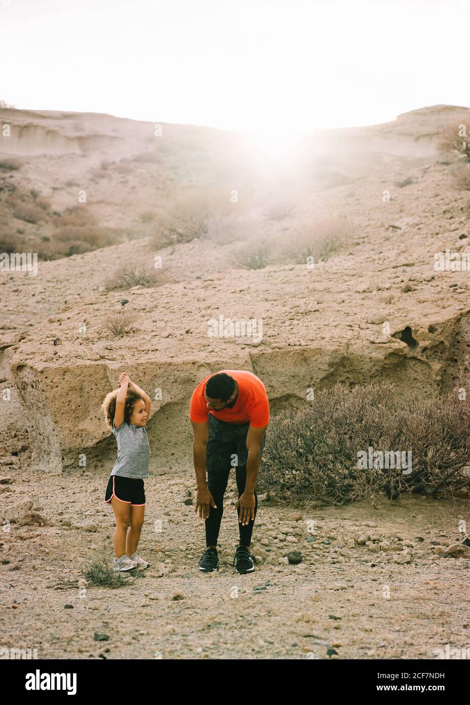 Vista laterale dell'uomo alato afroamericano in piedi activewear di fronte al bambino arricciato che insegna a stretching parte anteriore della coscia su sabbia orizzontale Foto Stock