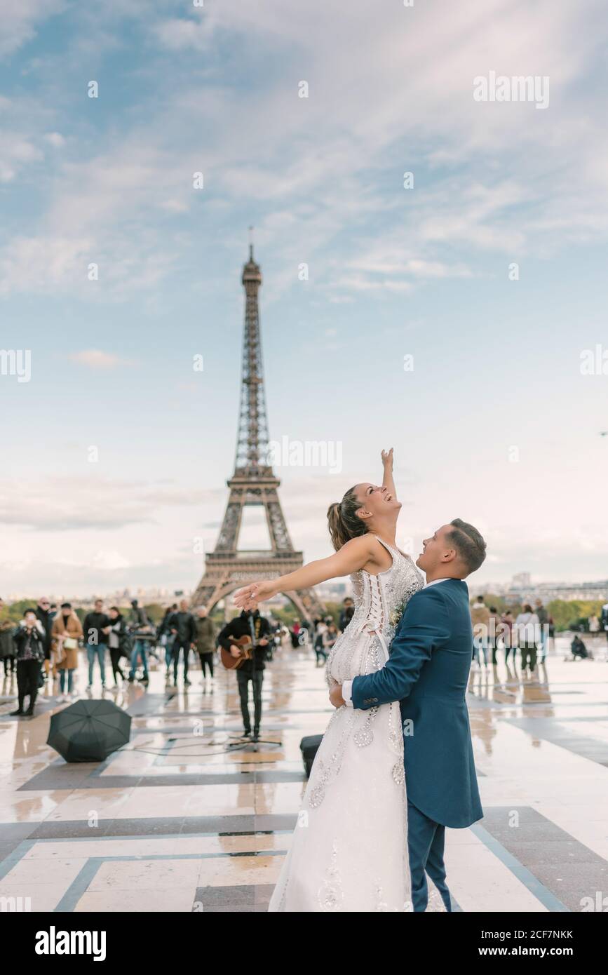 Sposo in tuta blu che solleva allegra sposa a braccia aperte In abito da sposa bianco con la Torre Eiffel sullo sfondo a. Parigi Foto Stock