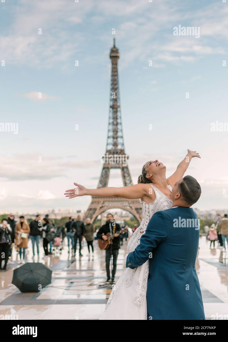 Sposo in tuta blu che solleva allegra sposa a braccia aperte In abito da sposa bianco con la Torre Eiffel sullo sfondo a. Parigi Foto Stock