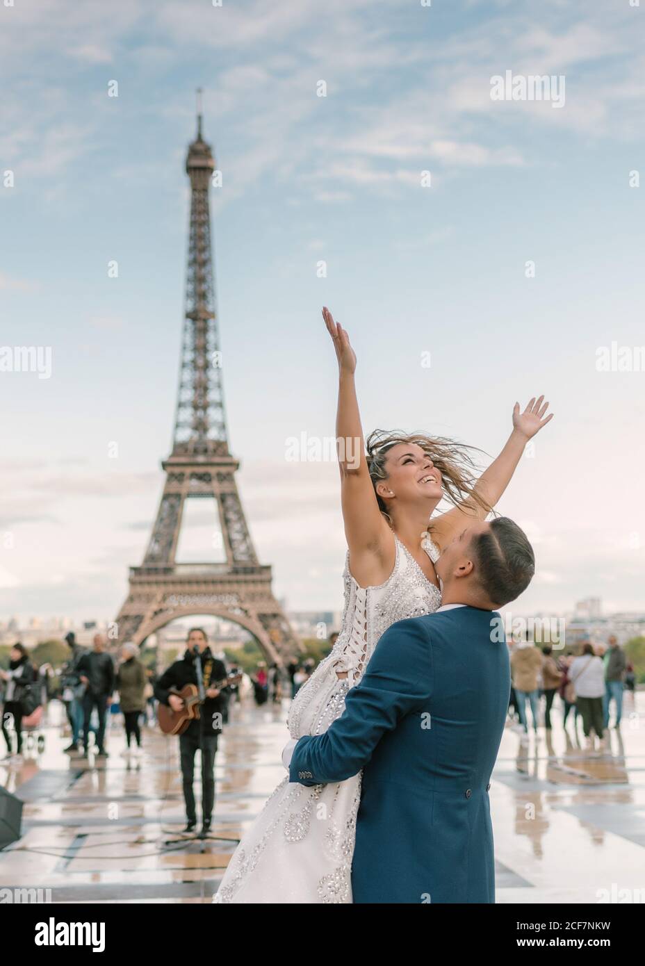 Sposo in tuta blu che solleva allegra sposa a braccia aperte In abito da sposa bianco con la Torre Eiffel sullo sfondo a. Parigi Foto Stock
