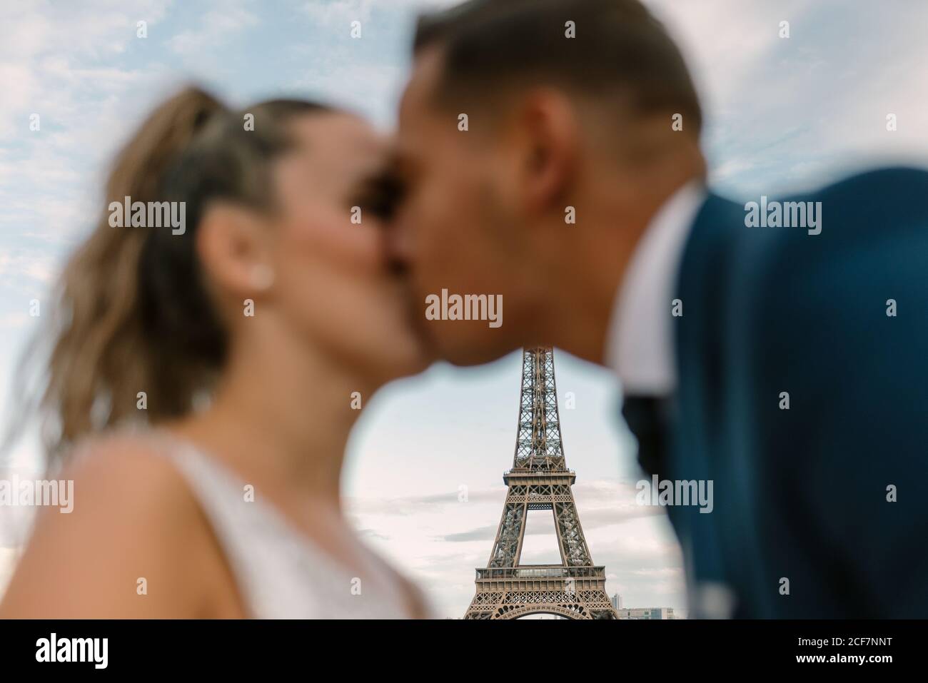 Groom fuori fuoco in tuta blu e sposa dentro Abito da sposa bianco baciando appassionatamente con la Torre Eiffel sullo sfondo A Parigi Foto Stock
