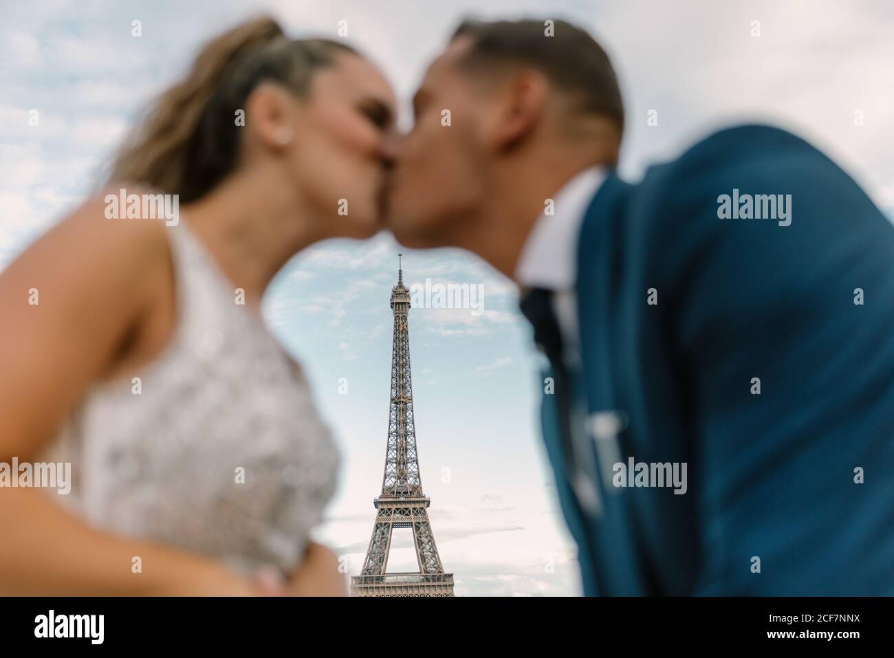 Groom fuori fuoco in tuta blu e sposa dentro Abito da sposa bianco baciando appassionatamente con la Torre Eiffel sullo sfondo A Parigi Foto Stock