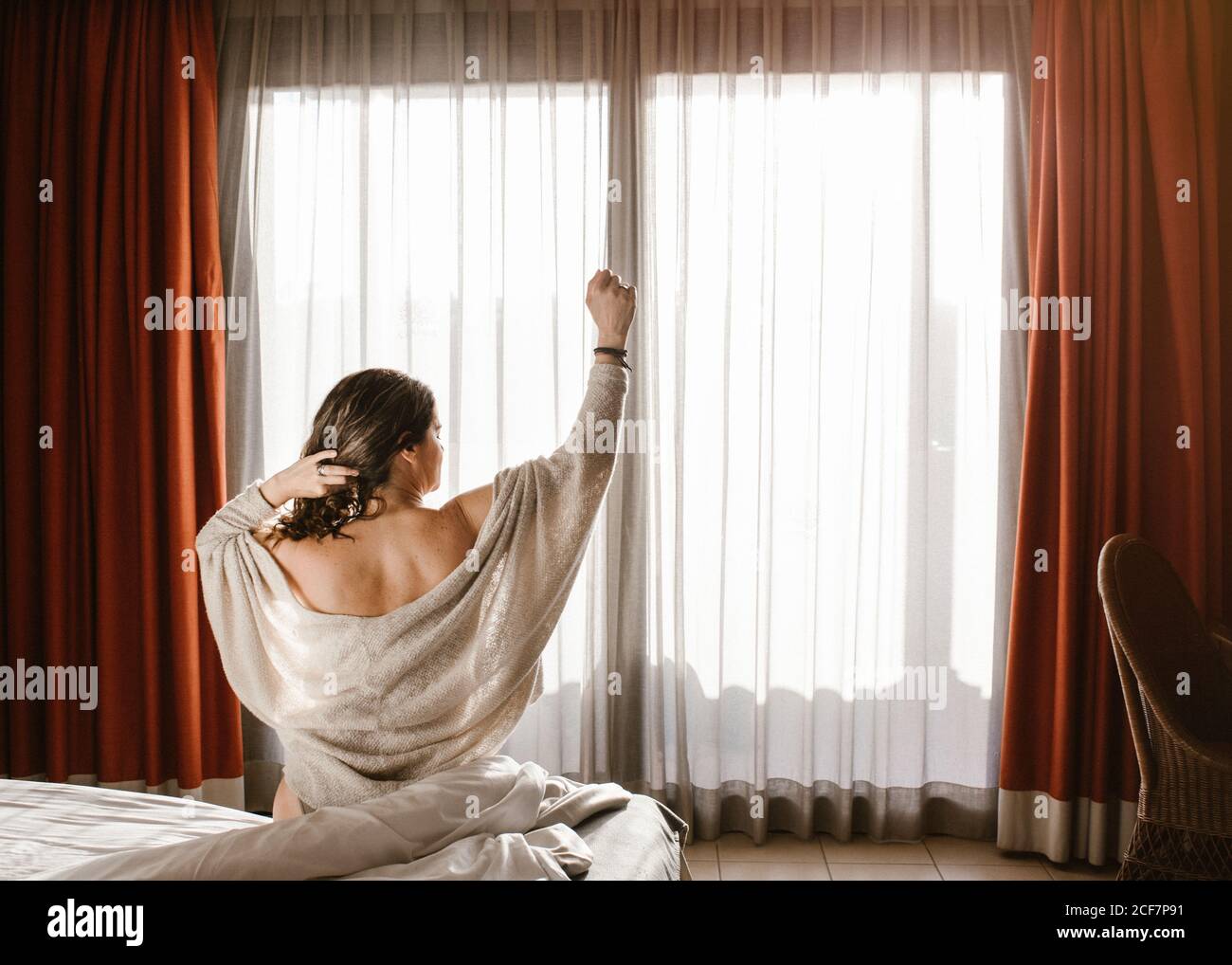 Vista posteriore della femmina adulta con braccio sollevato che guarda lontano e toccando i capelli mentre si siede sul letto contro la finestra chiusa con tende rosse e tulle bianco al mattino soleggiato Foto Stock