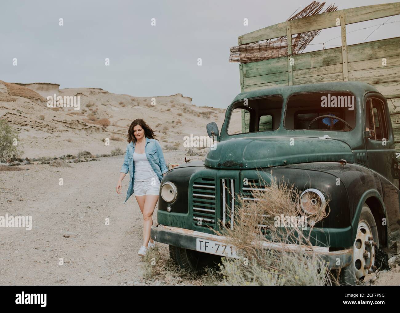 Giovane donna in pantaloncini e maglietta in denim camminando su camion verde con corpo in legno e guardando lontano sullo sfondo della natura Foto Stock