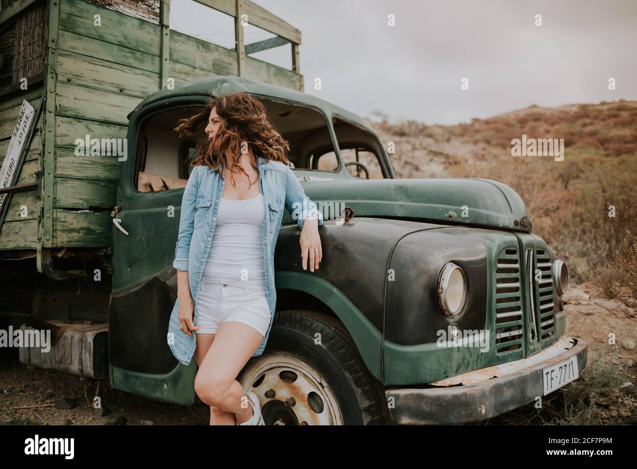 Giovane donna in pantaloncini e camicia in denim in piedi da camion verde con corpo in legno e guardando lontano sullo sfondo della natura Foto Stock