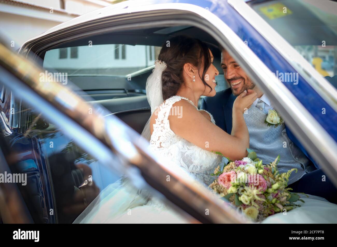 Sposa felice in abito da sposa e sposo in abito di classe seduto in un'automobile d'epoca e guardandosi l'un l'altro Foto Stock
