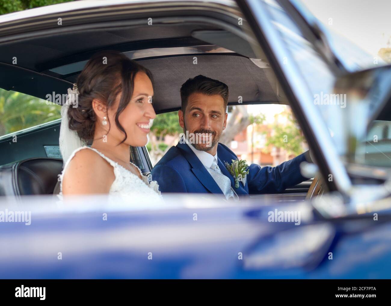 Sposa felice in abito da sposa e sposo in abito di classe seduto in un'automobile d'epoca e guardandosi l'un l'altro Foto Stock