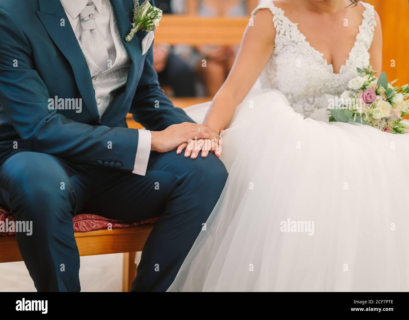 sposo irriconoscibile in abito elegante con boutonnière e sposa in abito da sposa bianco seduto sulla panca e tenendo le mani durante cerimonia in chiesa Foto Stock