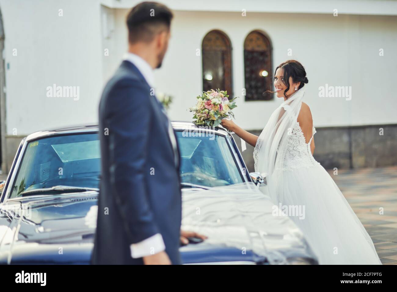Vista laterale di una sposa sorridente che indossa un elegante abito da sposa e. veil in piedi con bouquet vicino auto di lusso e guardando sposo in un abito elegante Foto Stock