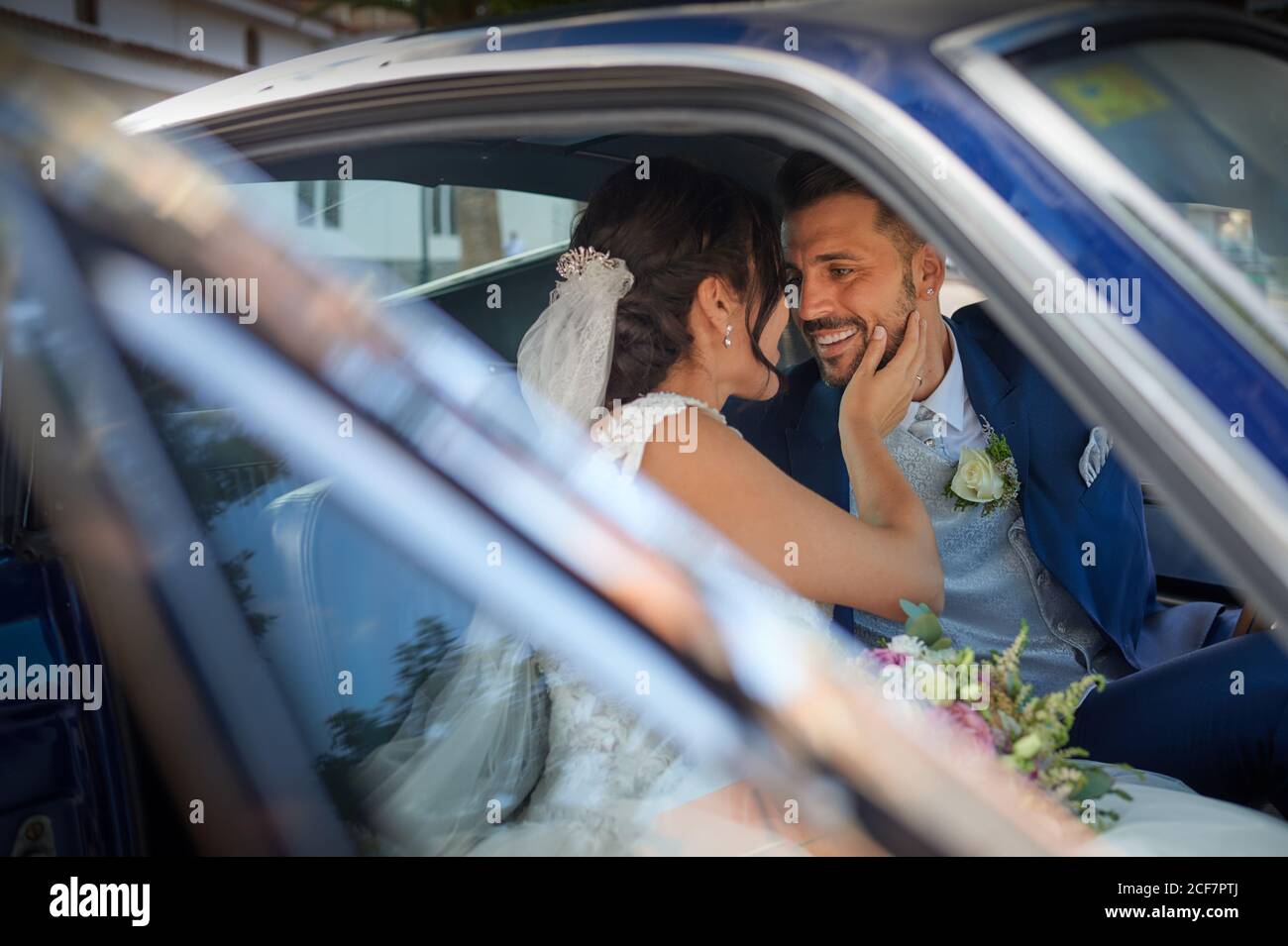 Sposa felice in abito da sposa e sposo in abito di classe seduto in un'automobile d'epoca e guardandosi l'un l'altro Foto Stock