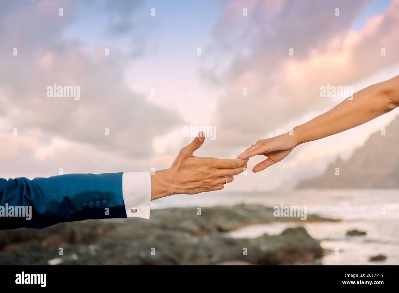 uno sposo irriconoscibile in un abito elegante e una sposa che tocca delicatamente le dita mentre si è in piedi sulla costa rocciosa durante il magnifico tramonto Foto Stock