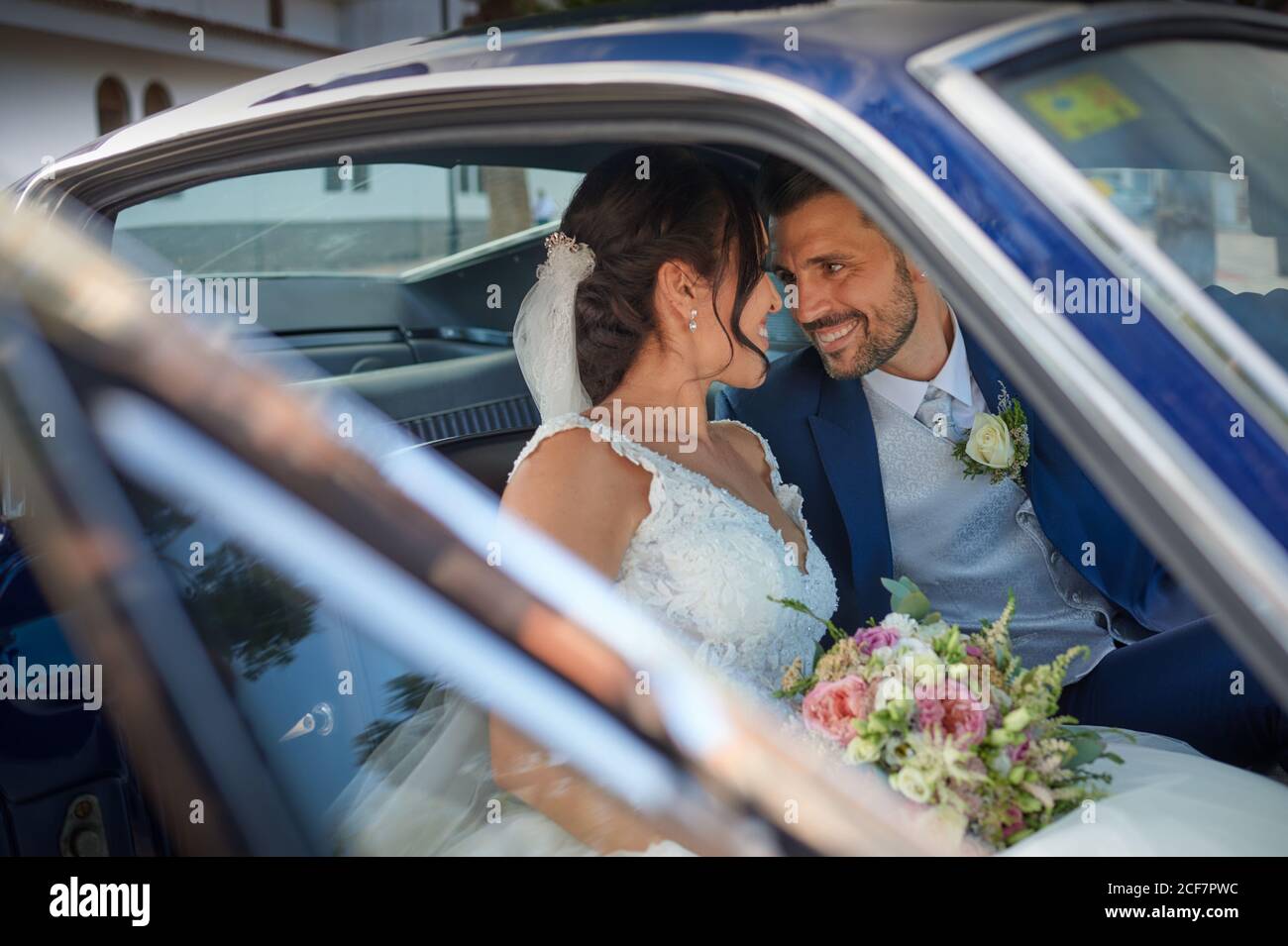 Sposa felice in abito da sposa e sposo in abito di classe seduto in un'automobile d'epoca e guardandosi l'un l'altro Foto Stock
