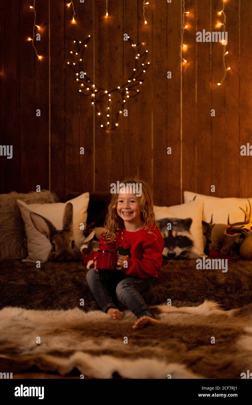Adorabile bambina che beve da una tazza rossa mentre si siede in una stanza piena di decorazione di natale Foto Stock