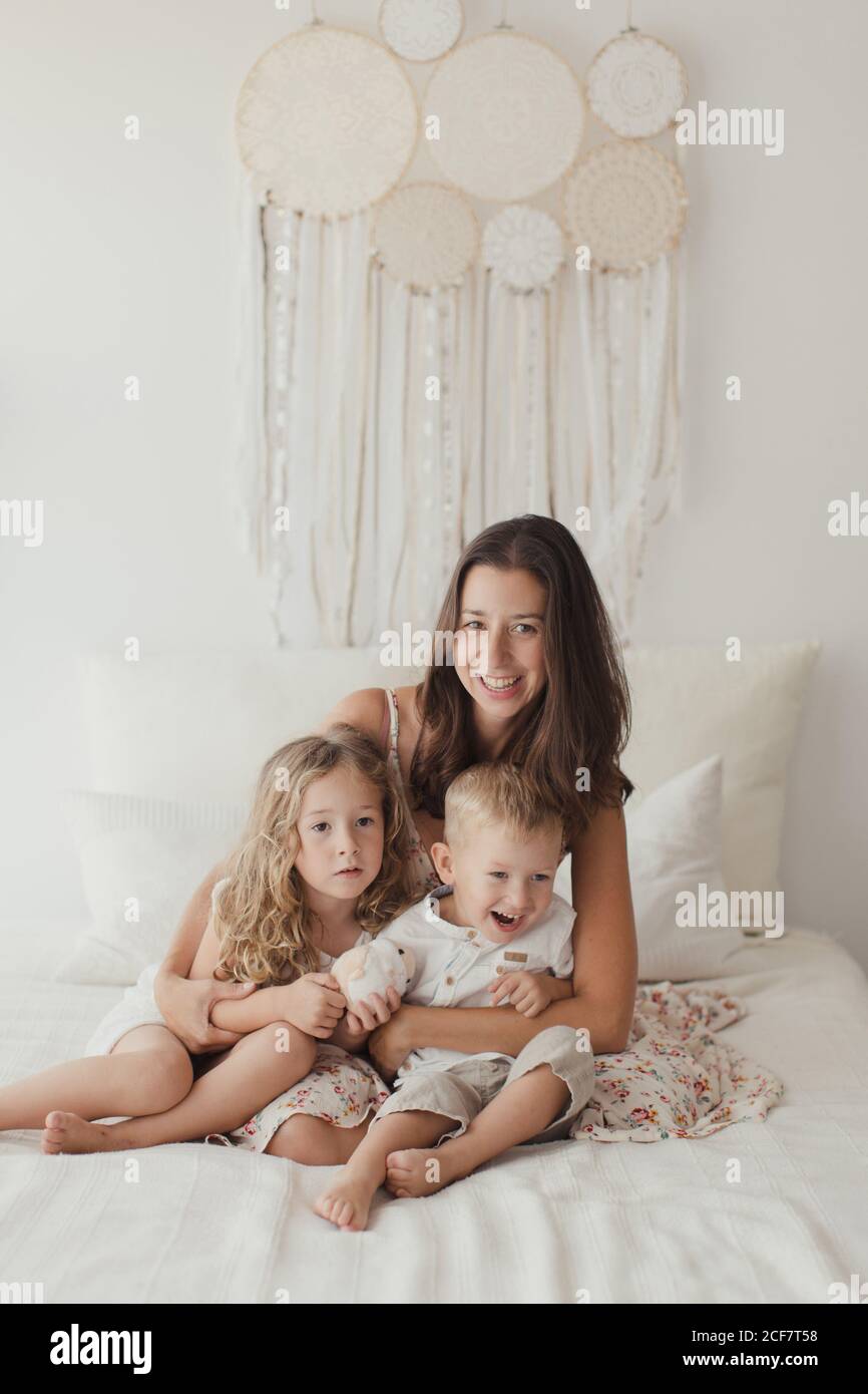 Una giovane brunetta positiva seduta sul letto e un piccolo maschio che si agganava e le donne bambini guardando la macchina fotografica in elegante camera da letto Foto Stock