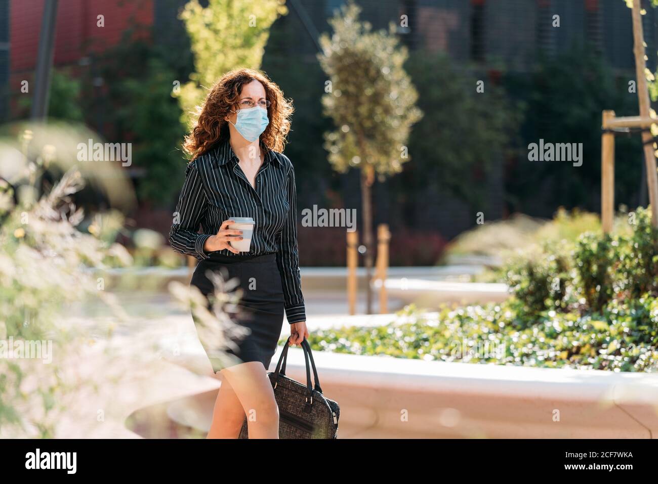 Donna che passa al suo lavoro indossando una maschera e tenendo una tazza di caffè mentre cammina attraverso un parco. Foto Stock