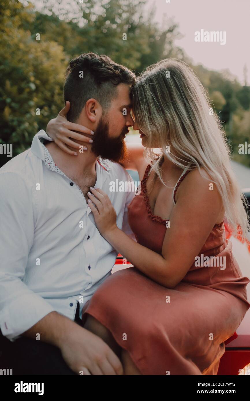 Tenero uomo barbuto in camicia bianca e pantaloncini neri che abbraccia sorridente Donna in vestito mentre si siede sul cappuccio rosso dell'automobile sull'autostrada vicino alla foresta verde Foto Stock