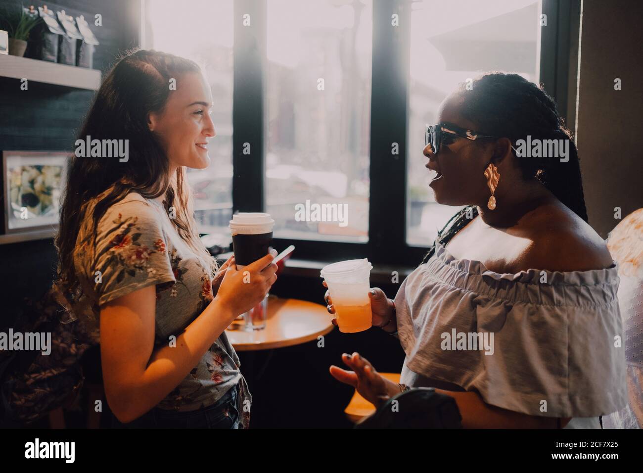 Vista laterale di allegre donne casual multirazziale e ridere bere caffè mentre si siede vicino alla finestra al caffè al tramonto Foto Stock