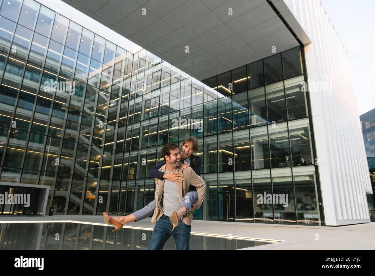 Felice uomo adulto in abiti casual trasportare Donna piggyback mentre si è in piedi e ridere sulla strada contro centro business contemporaneo in centro Foto Stock