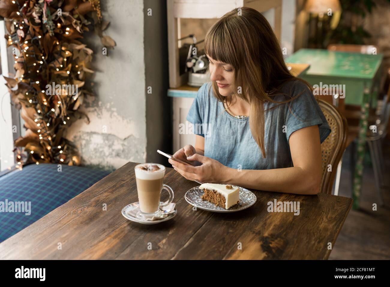 Bionda giovane allegra femmina in abiti casual digitando messaggio su smartphone mentre si siede a tavola con caffè e dessert caffè accogliente Foto Stock