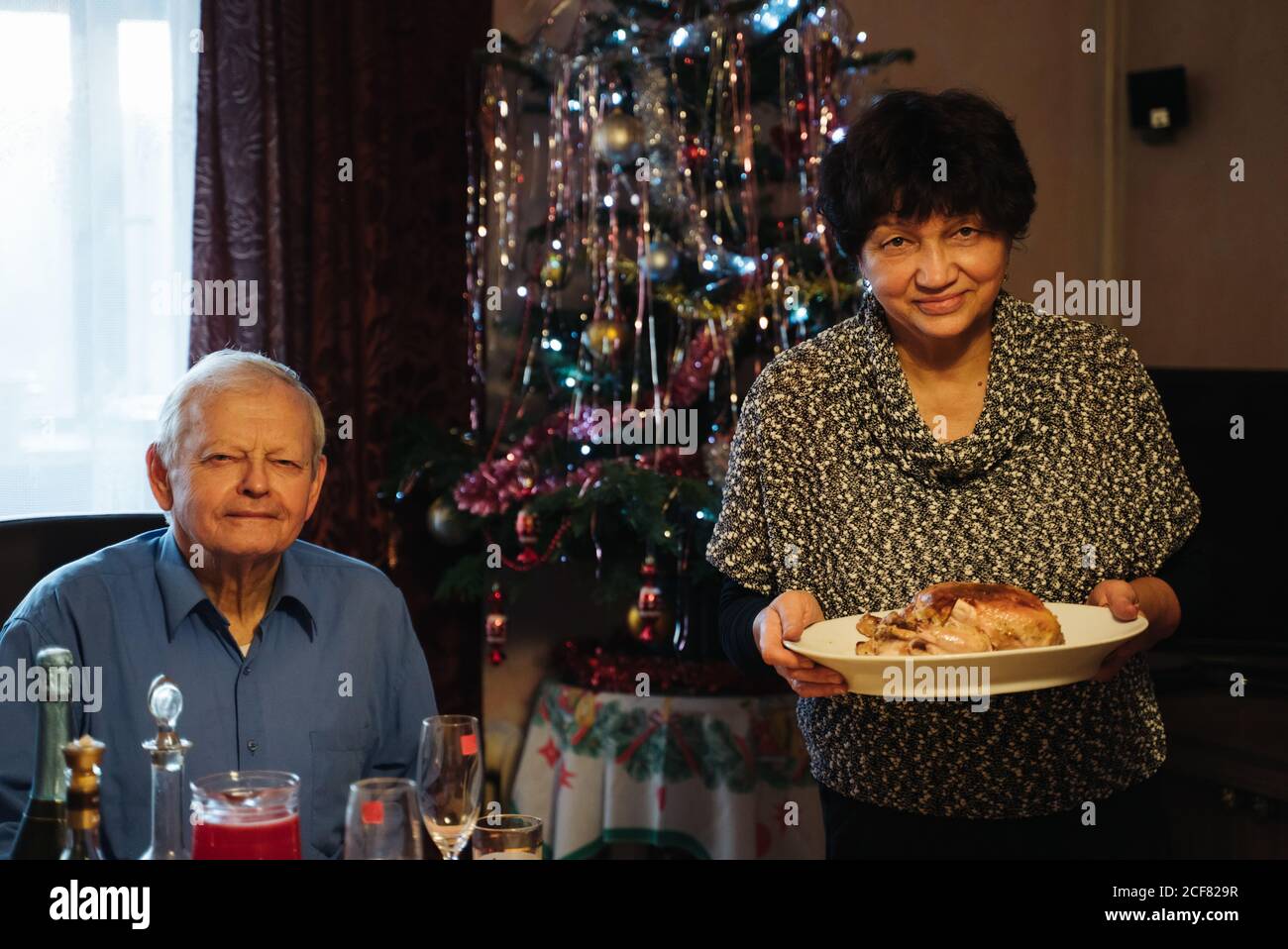 Positivo Senior Woman Holding piatto con pollo arrosto e sorridente marito seduto al tavolo durante la cena di famiglia a casa con albero di Natale in background Foto Stock
