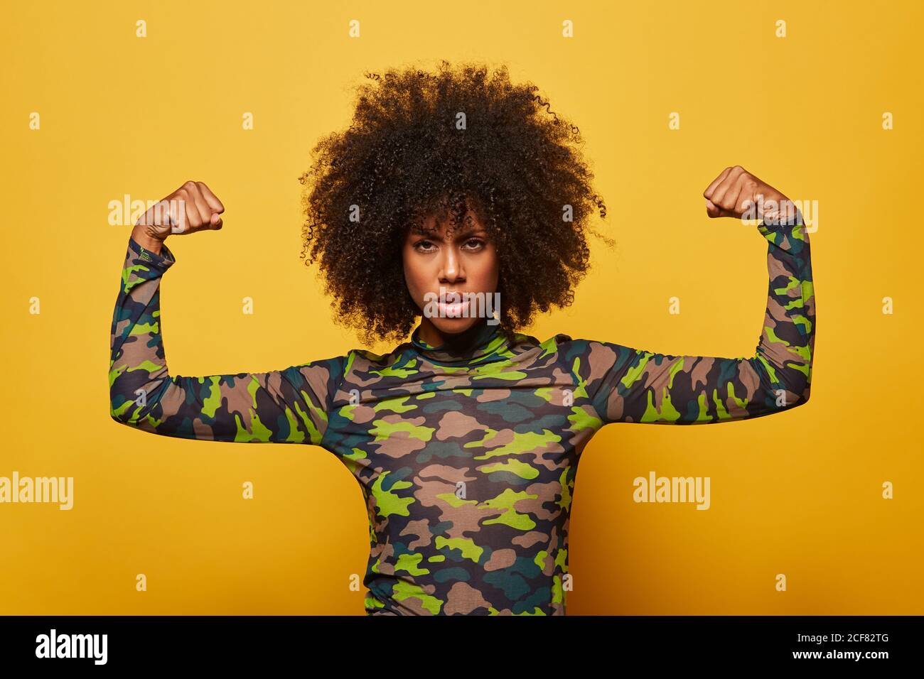 Seria forte etnia Empowered Donna in camicia mimetizzazione e con acconciatura afro mostrando bicipiti su sfondo giallo guardando la macchina fotografica Foto Stock