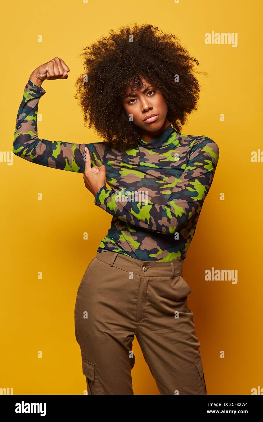 Seria forte etnia Empowered Donna in camicia mimetizzazione e con acconciatura afro mostrando bicipiti su sfondo giallo guardando la macchina fotografica Foto Stock