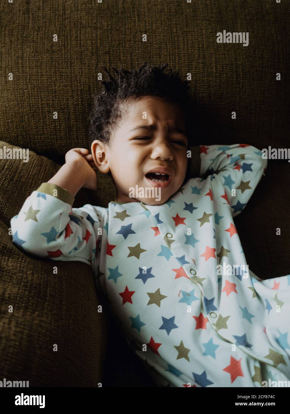 Carino afroamericano ragazzo in pigiama sdraiato su un comodo divano e iniziare a piangere a casa Foto Stock