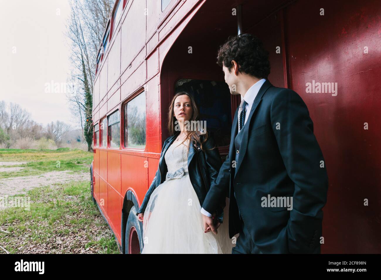 Sposa in abito da sposa e giacca e groom in pelle abito elegante che tiene le mani e si trova vicino a due piani shabby autobus mentre si guardano l'un l'altro Foto Stock
