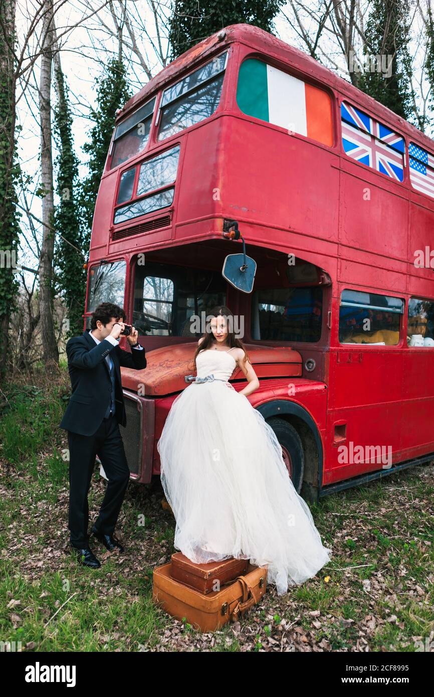 Elegante sposo in tuta scattando foto di sposa in bianco abito da sposa in piedi su valigie vintage vicino autobus a due piani Foto Stock