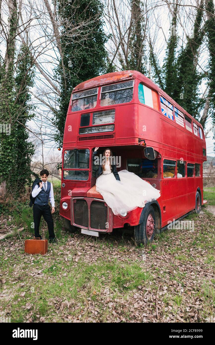 Sposa in abito da sposa seduta sul cappuccio della vecchia doppia autobus e sposo in elegante tuxedo in piedi vicino alla valigia in legno Foto Stock
