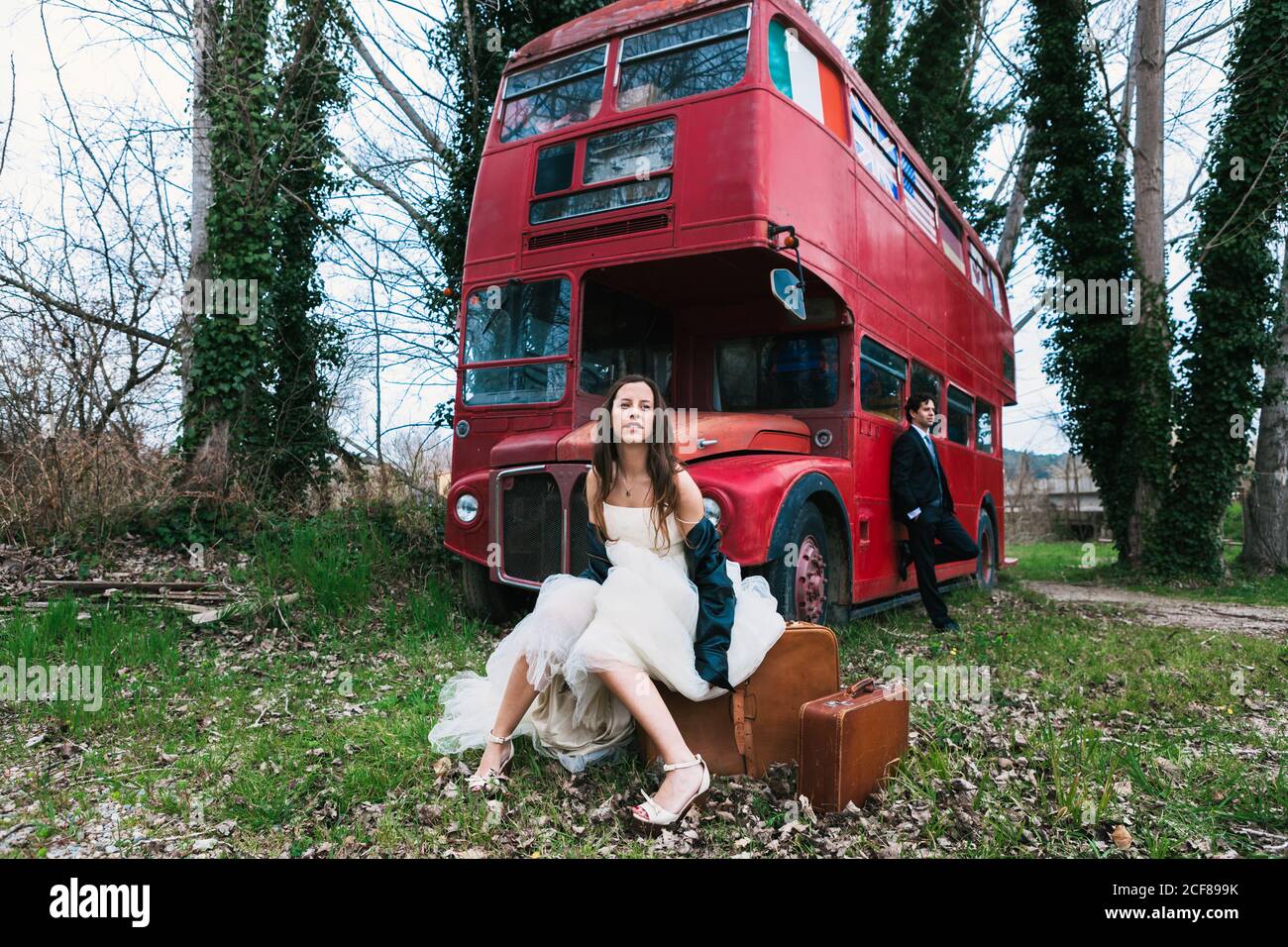 Sposa in abito da sposa e giacca in pelle seduta su retro valigia e sposo in tuta appoggiati su un doppio ponte rosso autobus abbandonato in foresta Foto Stock