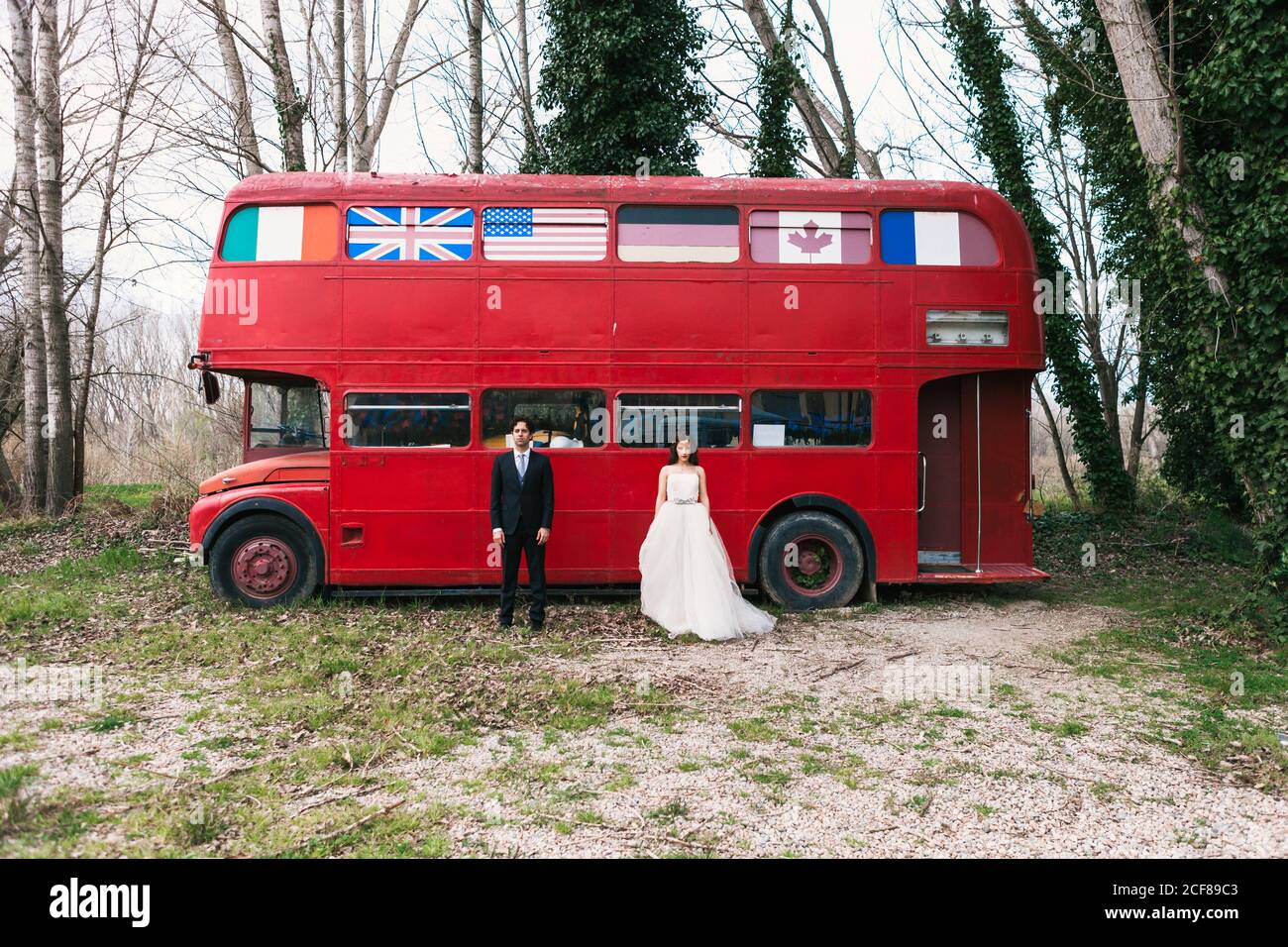 Sposa in elegante abito da sposa e sposo in abito di classe in piedi vicino al bus rosso vintage durante il giorno del matrimonio e cercando alla telecamera Foto Stock
