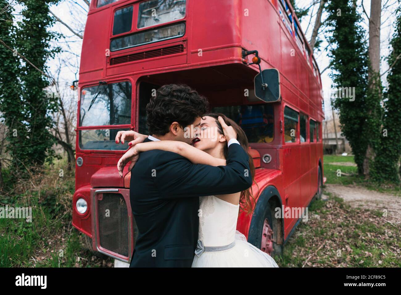 Vista laterale di sposa e sposo in amore abbracciando e. baciarsi nella foresta mentre si sta in piedi vicino all'autobus rosso retrò giorno di nozze Foto Stock