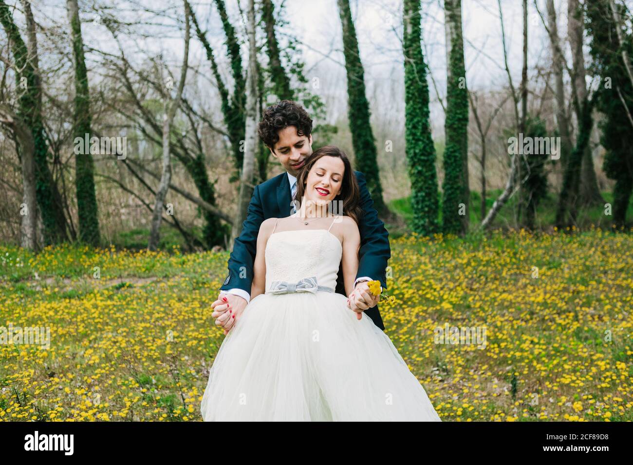 Sposo felice in abito elegante abbracciando sposa sorridente con il giallo dandelions su prato in fiore durante il giorno del matrimonio Foto Stock