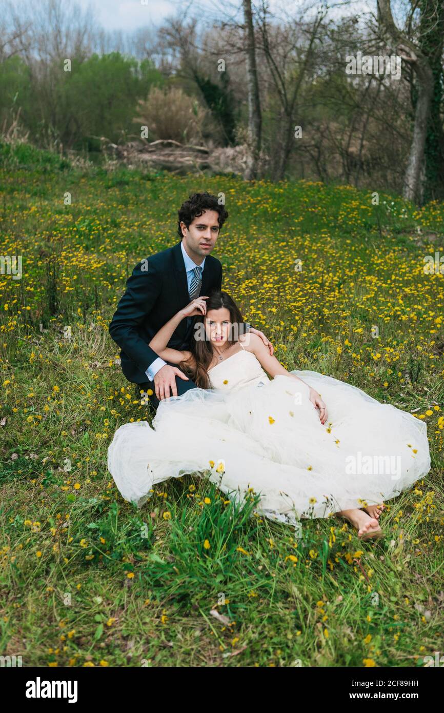 Alto angolo di stile calmo in tuta di classe e a piedi nudi sposa in abito da sposa che abbraccia in prato circondato da giallo fiori Foto Stock
