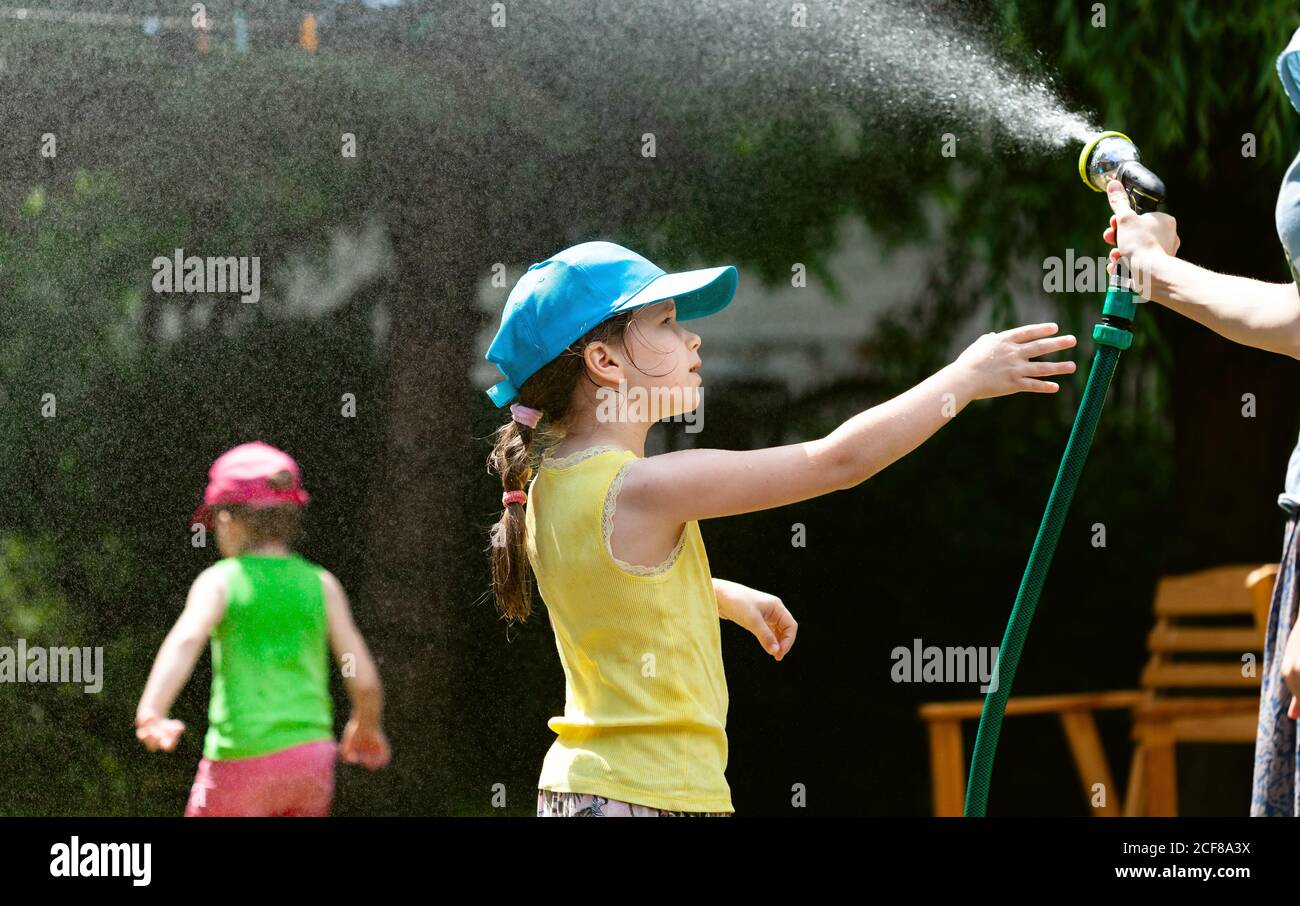 Bambini che giocano con il tubo dell'acqua nel giardino, madre che spruzzano l'acqua nell'aria, ragazza che raggiunge fuori per il tubo flessibile. Divertimento all'aperto per le famiglie Foto Stock