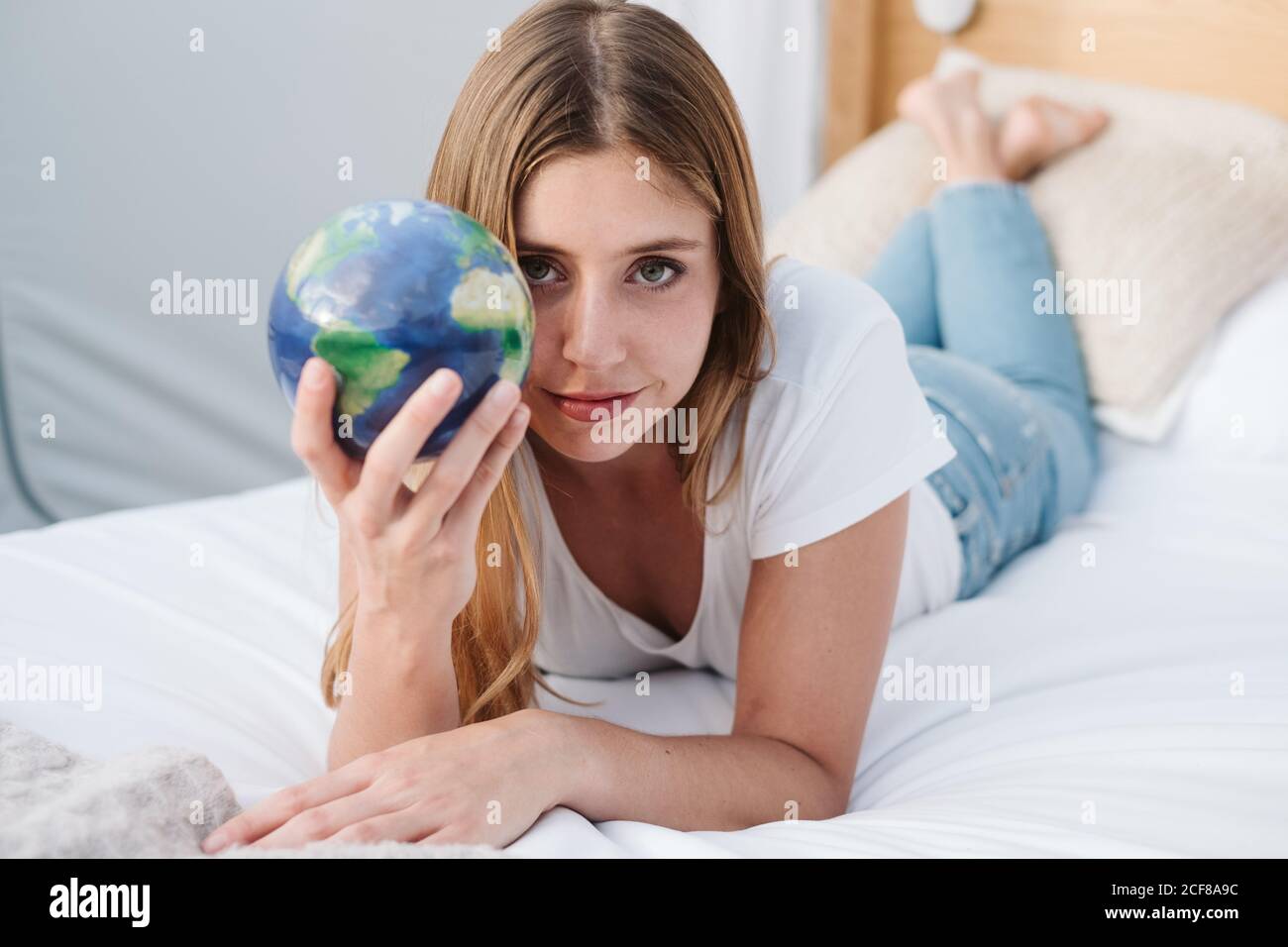 Bella donna in jeans e t-shirt bianca rilassante a letto con il globo in mano e guardando la macchina fotografica Foto Stock