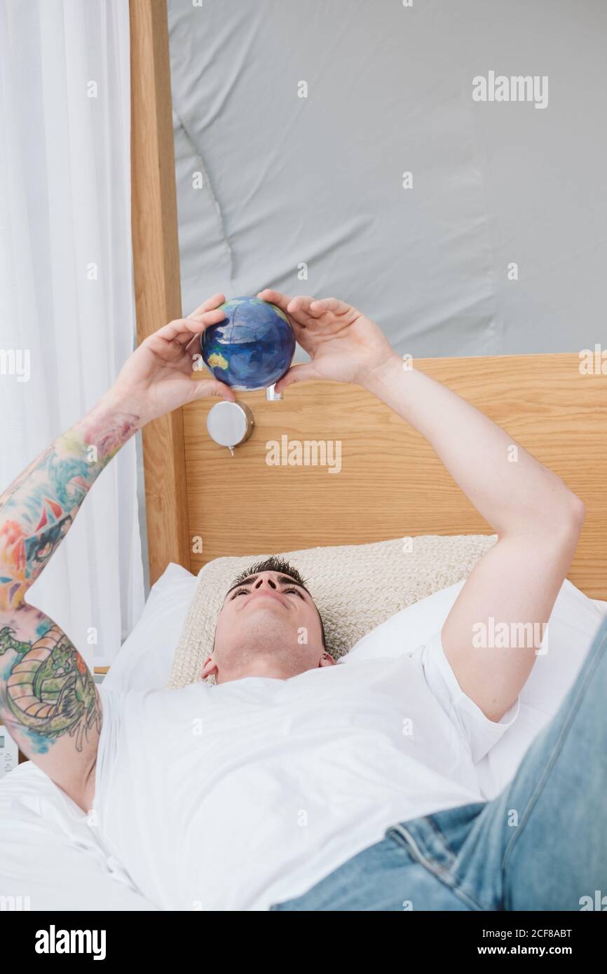 Bell'uomo tatuato in una maglietta bianca che si ritorce in mani piccole palla di terra mentre si rilassa a letto Foto Stock