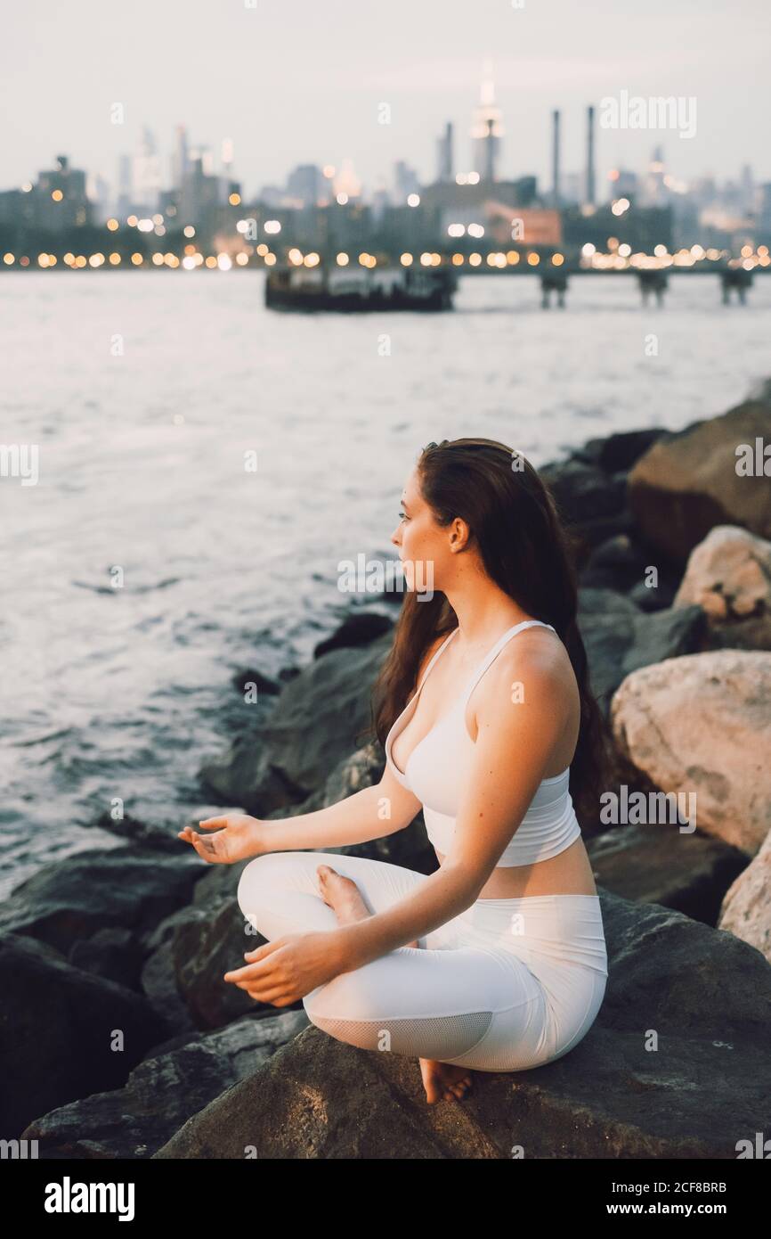 Vista dall'alto laterale di una femmina concentrata in attività seduta sulla pietra sul lungomare mentre si pratica yoga sullo sfondo di il paesaggio urbano serale e lo sguardo lontano Foto Stock